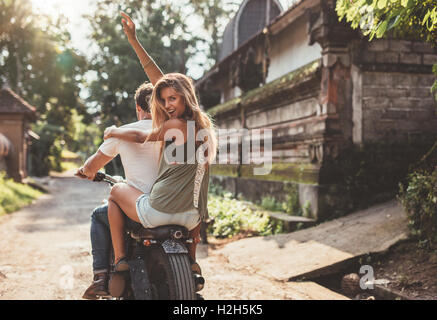Vue arrière shot of young couple tour de moto sur route du village. Belle jeune femme assise à l'arrière avec son bras rais Banque D'Images