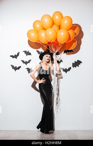 Toute la longueur de belle jeune femme en costume halloween sorcière orange holding balloons debout sur fond blanc Banque D'Images