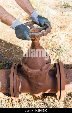 Gants de travail avec les mains serrant une valve du tuyau d'une très ancienne et ligne d'eau rouillée Banque D'Images