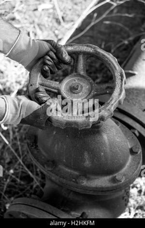 Gants de travail avec les mains serrant une valve du tuyau d'une très ancienne et ligne d'eau rouillée Banque D'Images