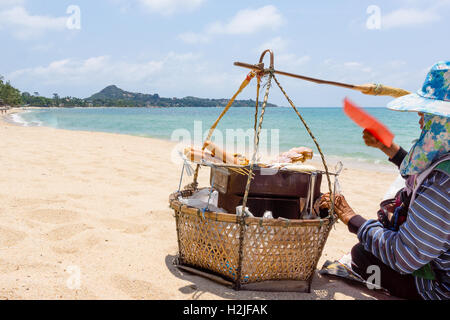 Vendeur thaï assis sur une plage de sable fin avec son panier à proximité de la mer sur le gril saucisses pour touristes Banque D'Images