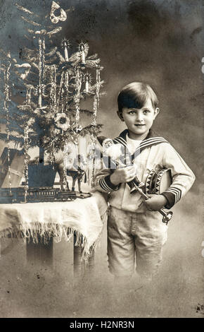 Heureux petit garçon avec arbre de Noël, cadeaux et jouets vintage. Photo sépia antique original avec du grain de film et des rayures Banque D'Images