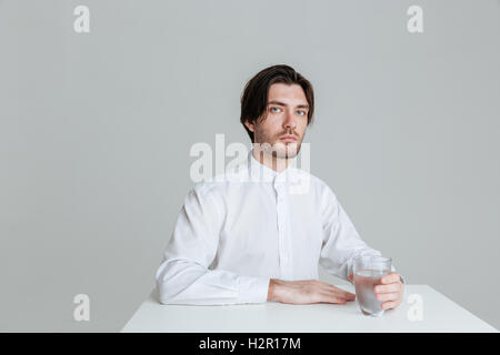Young l'homme assis à la table de verre d'eau isolé sur fond gris Banque D'Images