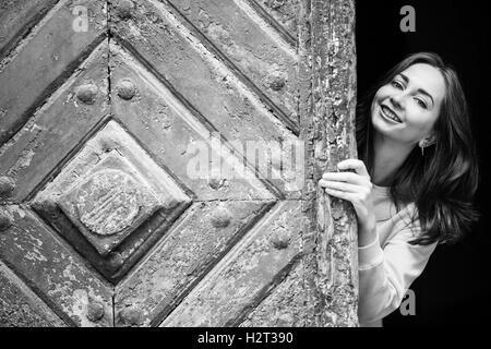 Jolie jeune fille de peering derrière l'ancienne porte en bois, en noir et blanc. Banque D'Images