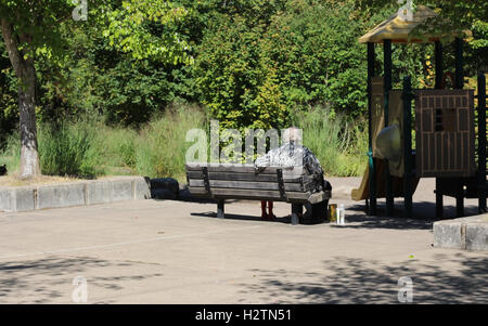 Femme âgée assise sur un banc dans le parc près de la structure de jeux pour les enfants, l'eau potable et manger des craquelins sur sunny summer Banque D'Images