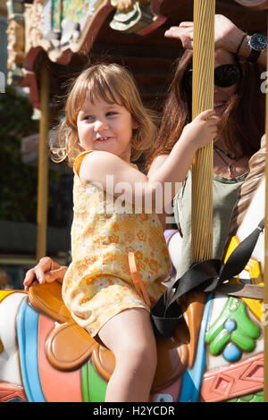 Portrait de trois ans blonde jolie fille avec robe jaune assis sur un des chevaux colorés dans un carrousel, à côté de brunette w Banque D'Images