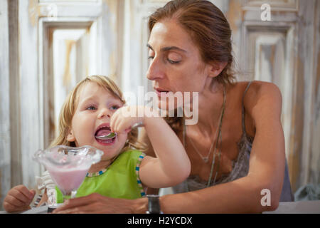 Trois ans avec enfant bib vert ouvrir grand la bouche eating strawberry ice cream avec cuillère à partir de la crystal cup à côté de mère à re Banque D'Images