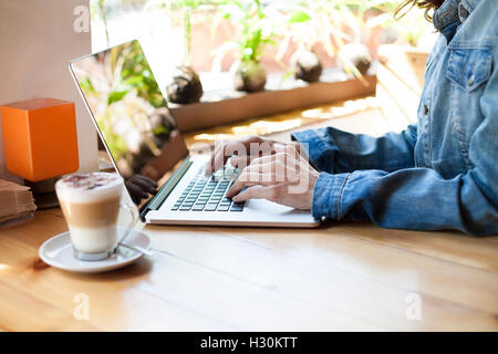 Veste jeans femme avec de taper au clavier de l'ordinateur portable à côté de la tasse de café cappuccino sur table en bois brun clair Banque D'Images