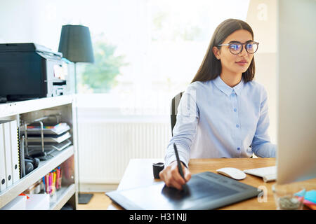 Young businesswoman travaillant dans un studio de design à l'aide d'une tablette électronique et comme elle travaille dans un grand ordinateur de bureau mo Banque D'Images