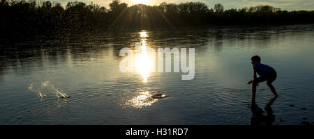 Garçon jette des pierres dans la rivière au coucher du soleil Banque D'Images