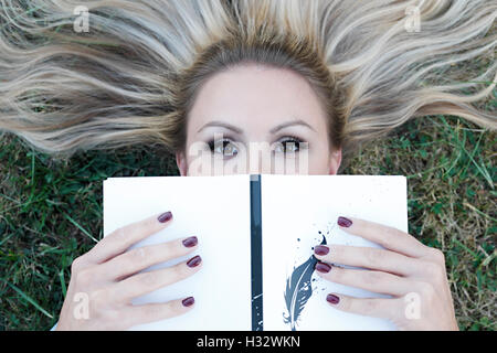 Belle jeune blonde allongée sur l'herbe avec un livre dans ses mains Banque D'Images