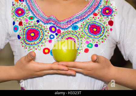 Libre young hispanic woman wearing blouse traditionnelle andine posant pour l'appareil photo holding green apple jusqu'en face de la poitrine, à l'intérieur de fruits du marché, la sélection d'aliments sains colorées en arrière-plan Banque D'Images