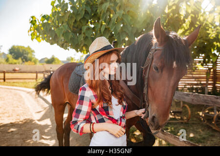 Belle jeune femme rousse cowgirl debout et prendre soin de son cheval dans le village Banque D'Images