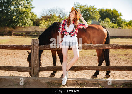 Toute la longueur de belle Rousse jeune femme cowgirl staning avec son cheval sur le ranch Banque D'Images