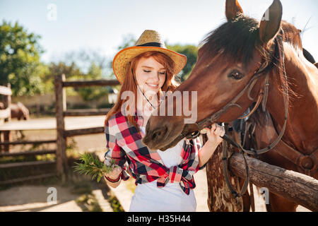 Cheerful rousse jeune femme cowgirl en donnant de la nourriture à cheval sur l'ranch Banque D'Images