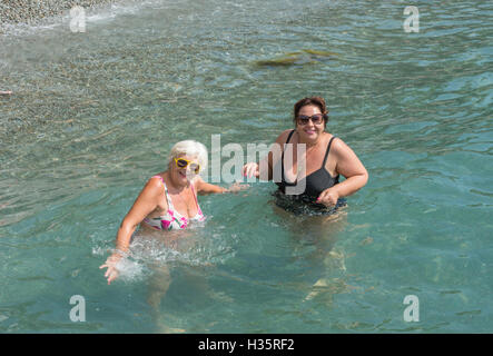 Deux femmes, blonde et brune dodue mature, sont debout dans l'eau clair transparent des galets de mer peu profonde. Banque D'Images