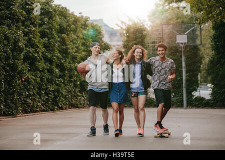 Longueur totale de la grenaille d'amis sur le terrain de basket-ball. Les jeunes hommes et femmes marchant avec skateboard et basketba Banque D'Images