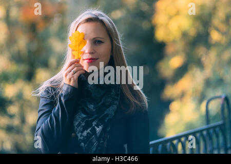 Belle jeune femme couvrant les yeux avec une feuille de chêne jaune contre l'arrière-plan d'automne colorés Banque D'Images