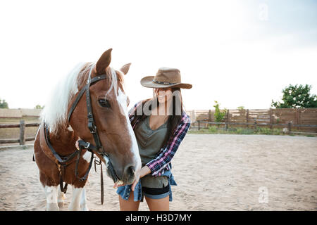 Cheerful young woman standing cowgirl et prendre soin de son cheval dans le village Banque D'Images