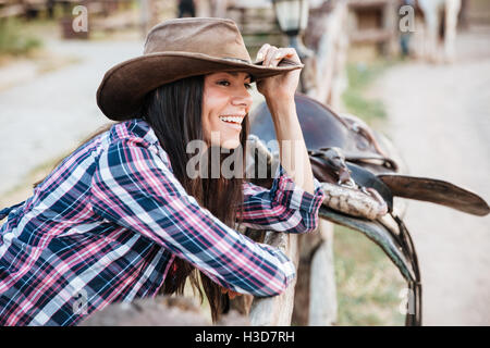 Smiling brunette jeune femme cowgirl leaning on fence in village Banque D'Images