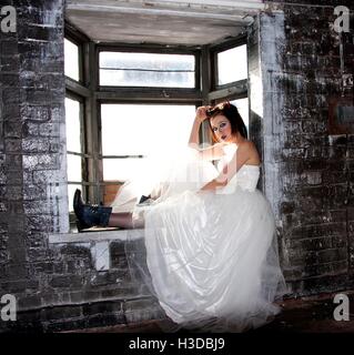 Young woman posing in bâtiment abandonné dans la fenêtre robe de mariage et des bottes noires Banque D'Images