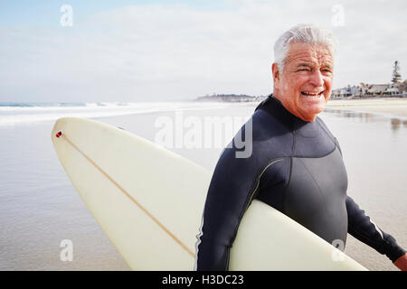 Smiling senior homme debout sur une plage, porter une combinaison isothermique et transporter une planche de surf.