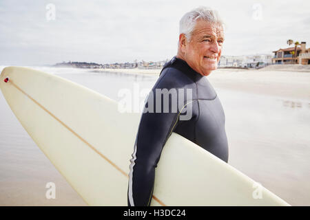 Smiling senior homme debout sur une plage, porter une combinaison isothermique et transporter une planche de surf.