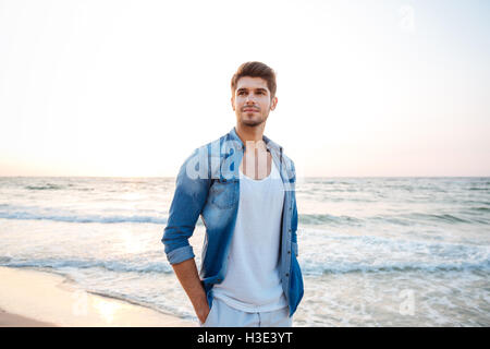 Beau jeune homme en jeans shirt debout sur la plage Banque D'Images