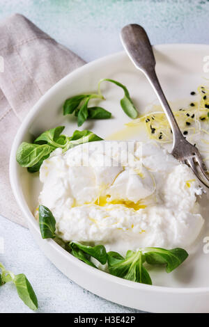 Tranches de fromage italien burrata avec fourche vintage, le maïs frais, salade de pousses et de l'huile d'olive dans une plaque en céramique blanche sur napki textiles Banque D'Images