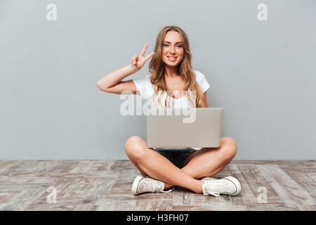 Smiling young woman with laptop sitting et montrant des signes de paix Banque D'Images