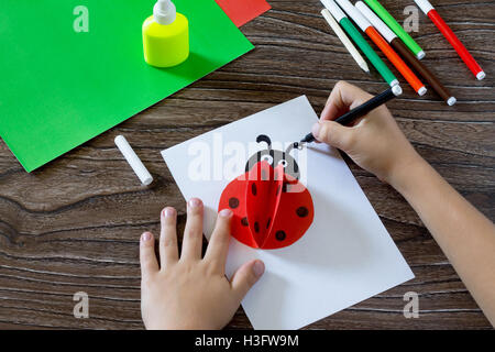 Attire l'enfant figurant sur une carte papier. L'enfant fait une carte postale avec coccinelle. Colle, papier, ciseaux sur une table en bois. Children's Banque D'Images