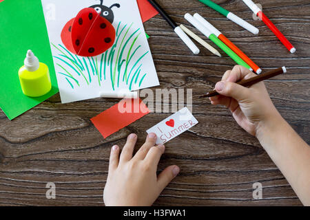 Document écrit que l'enfant détails J'adore l'été. L'enfant fait une carte postale avec coccinelle. Colle, papier, ciseaux sur un sol en bois t Banque D'Images