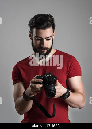 Jeune photographe barbu visualisation des photos sur l'écran LCD arrière de l'appareil photo reflex numérique. Moody désaturés portrait sur fond gris Banque D'Images