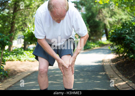 Closeup portrait, homme plus âgé en chemise blanche, pantalon gris, debout sur la route pavée, de graves douleurs au genou, d'arbres isolés à l'extérieur Banque D'Images