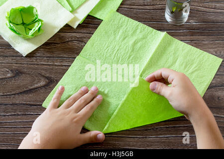 L'enfant est titulaire d'un carré de papier et pliez-le coin. L'enfant fait de l'artisanat de papier lily. Serviettes en papier sur une table en bois. Ch Banque D'Images