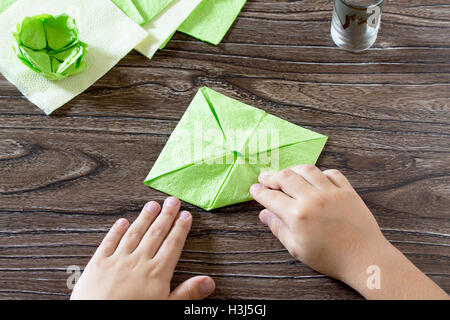 L'enfant est titulaire d'un carré de papier et pliez-le coin. L'enfant fait de l'artisanat de papier lily. Serviettes en papier sur une table en bois. Ch Banque D'Images