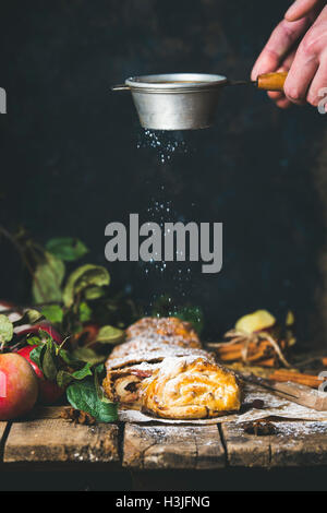 Strudel aux pommes gâteau coupé en morceaux avec des pommes rouges sur table en bois rustique et de l'homme ses mains avec de la poudre de sucre saupoudrer de grille Banque D'Images