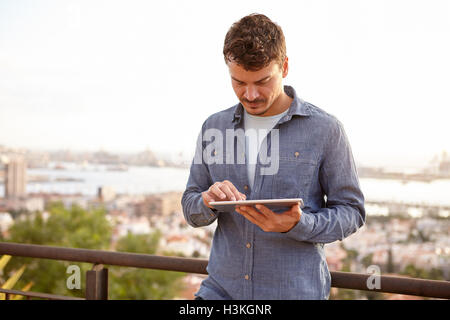 Mignon jeune homme attachant sur sa tablette portant une chemise en jean en étant debout sur un pont surplombant une ville Banque D'Images