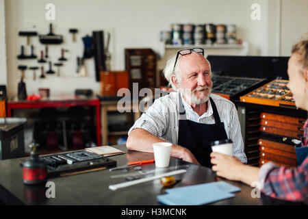 Artisan principal assis à table avec jeune artisane et souriant dans l'atelier des arts du livre Banque D'Images