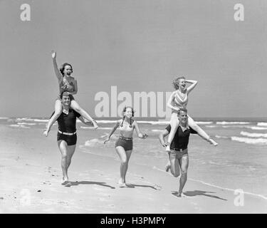 Groupe 1930 HOMMES FEMMES RUNNIG BATIFOLER SUR LA PLAGE À CHEVAL SUR LES ÉPAULES DES FEMMES POUR HOMMES Banque D'Images