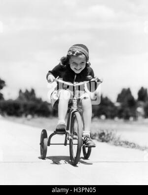 Années 1930 Années 1940 GIRL RIDING TRICYCLE SUR TROTTOIR Banque D'Images