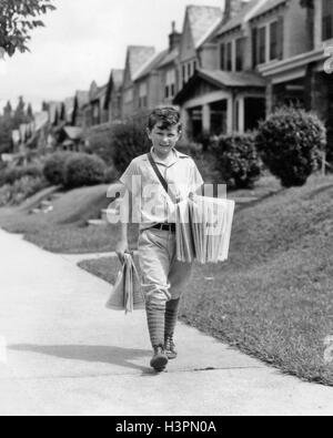 1930 garçon CRIEUR À LA CAMÉRA À LA LIVRAISON DE JOURNAUX À MARCHER LE LONG DE LA RUE DE BANLIEUE EN RANGÉE Banque D'Images