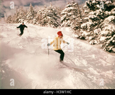 1970 DEUX PERSONNES HOMME FEMME SKI BAS pente enneigée PINS WOMAN WEARING YELLOW JACKET ET BONNET ROUGE Banque D'Images