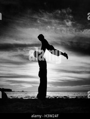 Années 1940 Années 1950 LA SILHOUETTE ANONYME HOMME PÈRE FILS ENFANT DE LEVAGE DANS L'AIR SUR LA PLAGE AU CRÉPUSCULE Banque D'Images