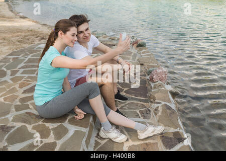 Couple aimant en tenant assis selfies au lac en ville Banque D'Images