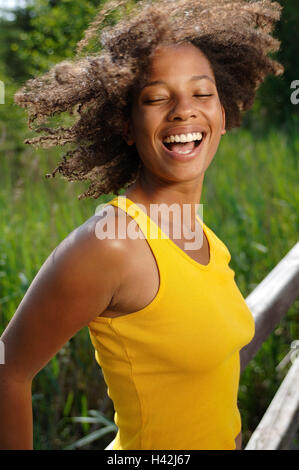 Jeune femme, rire, la peau sombre, pont, heureux, demi-portrait, les non-blancs, boucles, aux cheveux bruns, l'été, le soleil, profiter, loisirs, vacances, drôlement la vie, la joie, le plaisir, la joie, l'insouciance, de désinvolture, exhilarates, heureux, motion, top, débardeur, résumé, jaune, fondu, de la jeunesse, amuse, vivacité, vitalité, impulsivement, positivement, l'humeur, de transmission, de naturel, à l'extérieur de la jetée en bois, stand, mode de vie, Banque D'Images