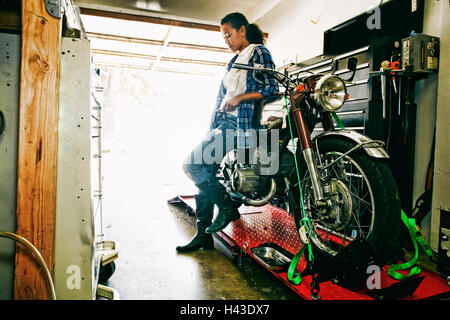 Mixed Race woman leaning on motorcycle in garage Banque D'Images