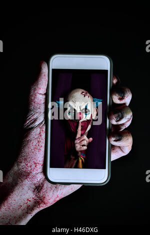 Libre d'un effrayant et sanglant main tenant un smartphone avec l'image d'un clown maléfique effrayant pour demander le silence dans son écran Banque D'Images