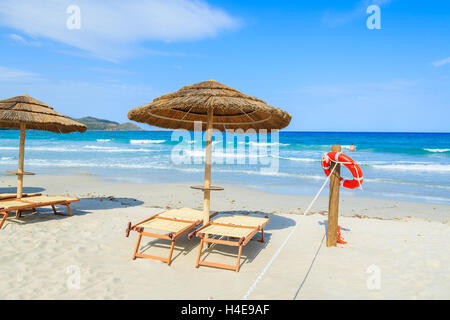 Chaises longues avec parasols et rouge bague la vie accrochée à un poteau en bois avec un jeune homme non identifié, la natation dans la mer d'azur de l'eau dans l'arrière-plan, l'île de Sardaigne, Italie Banque D'Images