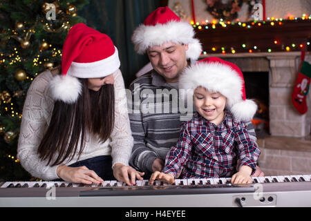 Famille - mère, père et enfant wearing santa hats jouant du piano sur fond de Noël Banque D'Images